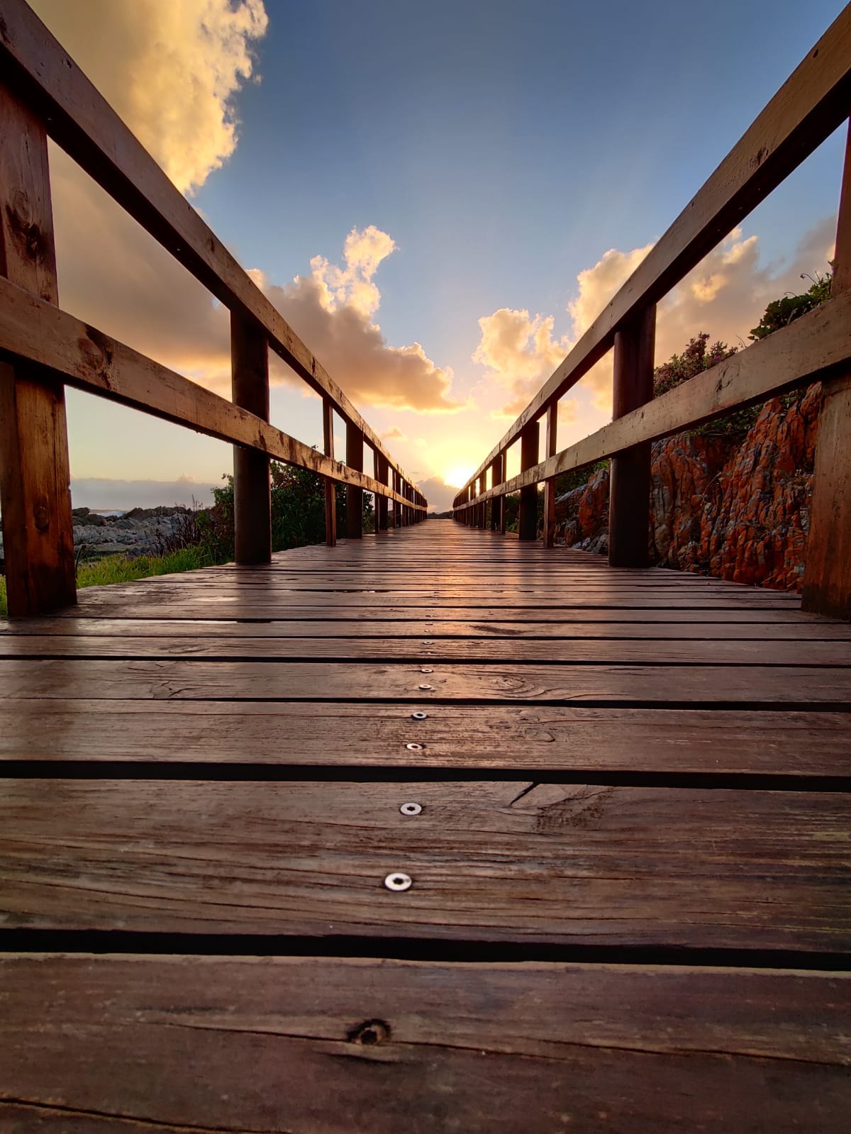 A timber boardwalk leading toward the sea at dusk beneath a tall cloudscape